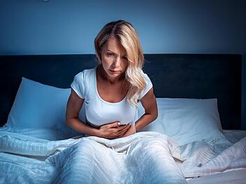 Darmgesundheit, eine blonde frau liegt im bett und kann nicht schlafen, sie hat schmerzen im gesicht und hält sich den Bauch, das zimmer ist dunkel