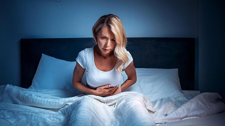 Darmgesundheit, eine blonde frau liegt im bett und kann nicht schlafen, sie hat schmerzen im gesicht und hält sich den Bauch, das zimmer ist dunkel