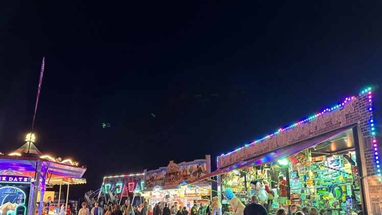 Ein nächtlicher Jahrmarkt mit bunten Lichtern, Buden und Fahrgeschäften. Menschen spazieren und genießen die festliche Atmosphäre der Gaustadter Kirchweih.