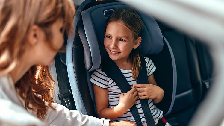 Eine nette Mutter setzt ihre Tochter in einen Autositz und schnallt sie an. Schutz während der Autofahrt.   A cute woman mother put her daughter in a car seat and fastens her seat belts. Protection during the trip in the car.