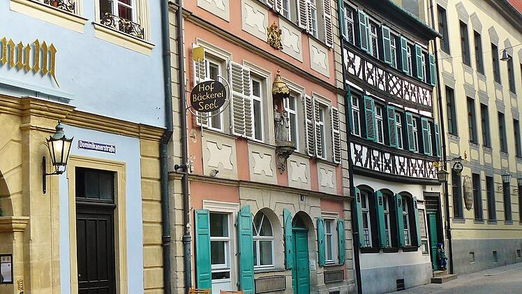 Bunte Altstadtfassaden mit Fachwerk und Au&szlig;enbestuhlung in einer ruhigen Stra&szlig;e. Ein Schild weist auf eine Hofb&auml;ckerei hin. Die Szene vermittelt eine charmante Atmosph&auml;re f&uuml;r einen Kurzurlaub.