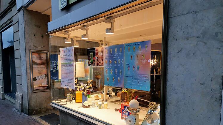 Ein Schaufenster in einer Stadt zeigt bunte Plakate mit Getränken und diverse Deko, passend zur Schließung eines Teeladens.