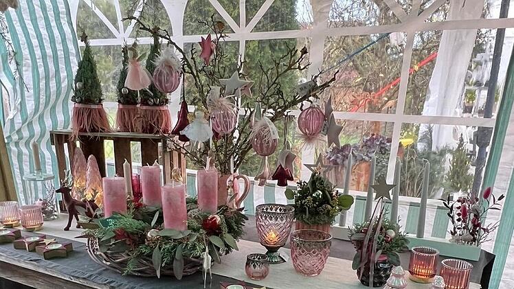 Ein weihnachtlich geschmückter Tisch mit rosa Kerzen, Sternen, Zweigen und Dekorationen. Ein Adventskranz und Kerzenhalter runden die festliche Szenerie ab.