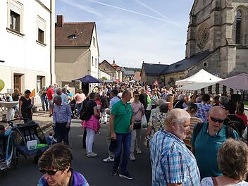 Viele Menschen besuchen eine geschäftige Straße bei sonnigem Wetter. Verkaufsstände und bunte Ballons sind zu sehen, ein reges Treiben herrscht. Im Hintergrund steht eine Kirche.