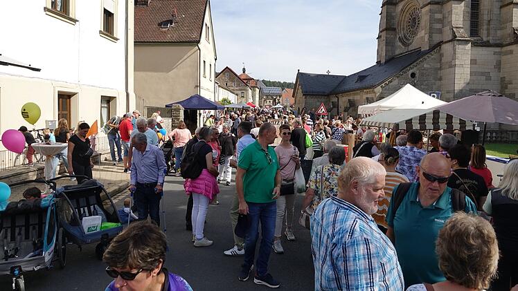 Viele Menschen besuchen eine geschäftige Straße bei sonnigem Wetter. Verkaufsstände und bunte Ballons sind zu sehen, ein reges Treiben herrscht. Im Hintergrund steht eine Kirche.