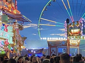 Ein bunt beleuchteter Jahrmarkt bei Nacht mit zahlreichen Besuchern, bunten Ständen und einem riesigen Riesenrad im Hintergrund. Große Menschenmengen genießen die festliche Atmosphäre.