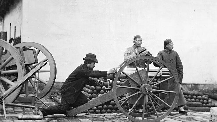 Aus der Zeit der Opiumkriege: John Thomson (links) posiert an einer Gatling-Kanone, bereit die Waffe abzufeuern. Neben ihm stehen zwei chinesische M&auml;nner, von denen einer den Verschluss der Kanone h&auml;lt.