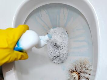 Eine weibliche Hand in einem gelben Gummihandschuh reinigt die Toilettensch&uuml;ssel mit einer B&uuml;rste und bedeutet  - Female hand in a yellow rubber glove cleans the toilet bowl with a brush and means
