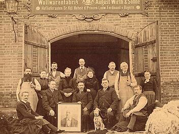 Die Familie Wirth (Bildmitte) mit ihren Webern und Spinnerinnen vor dem Firmengebäude an der Schütt (heute Versicherungsbüro Wirth und Firma Kuwe) mit einem Bild des Firmengründers. Rechts im Bild liegt ein Ballen Schafwolle zum Zerkleinern (Kartätschen). Foto: Familie Wirth