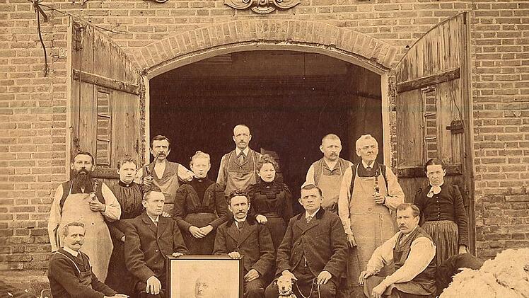 Die Familie Wirth (Bildmitte) mit ihren Webern und Spinnerinnen vor dem Firmengebäude an der Schütt (heute Versicherungsbüro Wirth und Firma Kuwe) mit einem Bild des Firmengründers. Rechts im Bild liegt ein Ballen Schafwolle zum Zerkleinern (Kartätschen). Foto: Familie Wirth