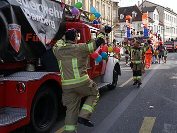 Ein Festumzug mit einem Feuerwehrwagen, geschm&uuml;ckt mit bunten Ballons. Feuerwehrleute in Kost&uuml;men feiern auf der Stra&szlig;e, umgeben von Zuschauern.