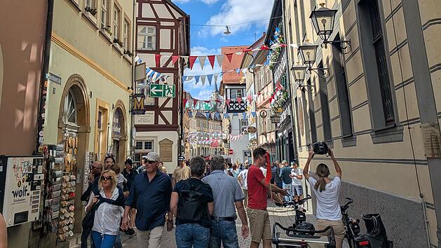 Bunt geschm&uuml;ckte Altstadtgasse mit fr&ouml;hlichen Menschen bei der Sandkerwa in Bamberg. F&auml;hnchen und Dekorationen erhellen die Stadt, w&auml;hrend Besucher entlang historischer Fassaden schlendern.