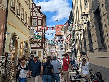 Bunt geschmückte Altstadtgasse mit fröhlichen Menschen bei der Sandkerwa in Bamberg. Fähnchen und Dekorationen erhellen die Stadt, während Besucher entlang historischer Fassaden schlendern.