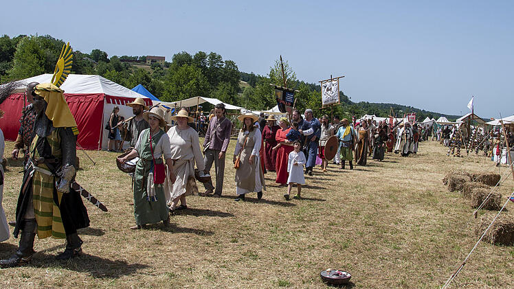 Menschen in mittelalterlichen Gewändern gehen auf einem Feld zwischen Zelten umher. Einige tragen Schilde und Fahnen. Die Szene erinnert an ein historisches Lager mit Pilgeratmosphäre.