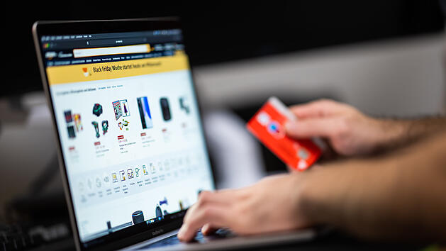 Ein Mann mit einer Bankkarte in der Hand sitzt am Laptop und schaut sich die Black Friday Angebote auf der Online-Shopping-Seite von Amazon an.