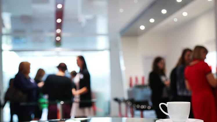 Eine weiße Tasse im Vordergrund, verschwommene Gruppe von Menschen im Hintergrund bei einer Veranstaltung oder Konferenz in einem modernen Raum.