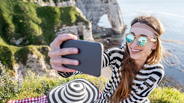 Young woman making selfie portrait with phone during the picnic outdoors sitting on the beautiful rocky coastline in France Junge Frau macht Selfie Porträt mit Telefon während des Picknick im Freien sitzen auf der schönen felsigen Küste in Frankreich