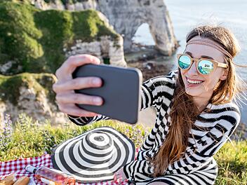 Young woman making selfie portrait with phone during the picnic outdoors sitting on the beautiful rocky coastline in France Junge Frau macht Selfie Porträt mit Telefon während des Picknick im Freien sitzen auf der schönen felsigen Küste in Frankreich