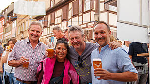 Menschen feiern auf der Bamberger Sandkerwa, l&auml;chelnd mit Biergl&auml;sern in der Hand, umgeben von Fachwerkgeb&auml;uden.
