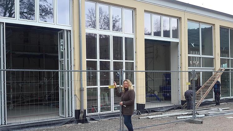 Kurdirektorin Andrea Schallenkammer zeigt den markanten Unterschied. Links im Bild sind bereits die neu eingebauten Ziehharmonika-Türen und die attraktiven Sprossenfenster in Nostalgie-Optik zu sehen. Im Hintergrund rechts bereiten Arbeiter gerade den Austausch weiterer alter Scheiben vor. Foto: Rolf Pralle