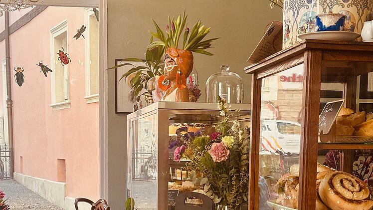 Gem&uuml;tliches Caf&eacute; mit Holzboden und Glasvitrine voller Geb&auml;ck. Blumendekoration und Pflanzen schaffen eine einladende Atmosph&auml;re. Ein neuer Anfang f&uuml;r Katharina H&uuml;ckst&auml;dt.