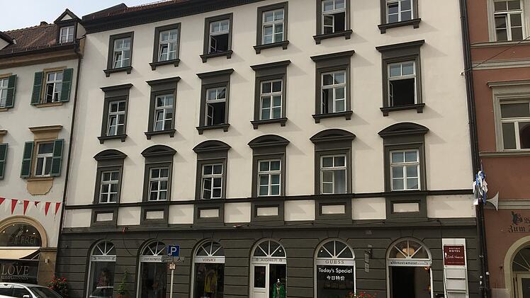 Historisches Gebäude mit drei Etagen und Geschäften im Erdgeschoss. Fahrräder und Autos stehen in der gepflasterten Straße. Oben hängt eine Girlande mit roten und weißen Wimpeln.