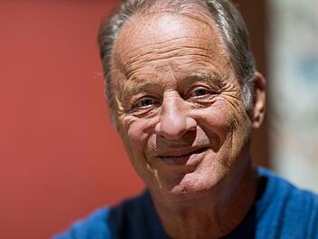 Ein &auml;lterer Mann mit grauen Haaren l&auml;chelt in die Kamera. Der Hintergrund ist unscharf und r&ouml;tlich gehalten.