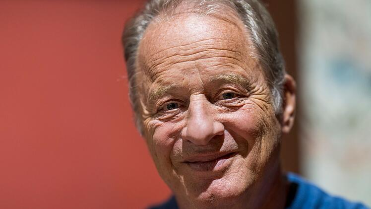 Ein &auml;lterer Mann mit grauen Haaren l&auml;chelt in die Kamera. Der Hintergrund ist unscharf und r&ouml;tlich gehalten.