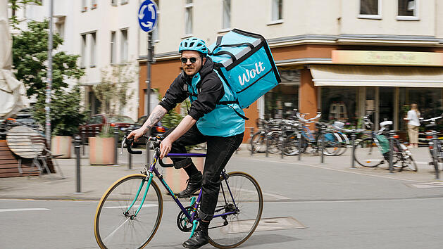 Ein Kurier auf einem Fahrrad tr&auml;gt eine gro&szlig;e, blaue Wolt-Liefertasche. Er f&auml;hrt durch eine st&auml;dtische Umgebung mit mehreren geparkten Fahrr&auml;dern im Hintergrund.