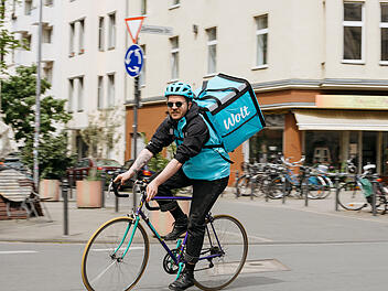 Ein Kurier auf einem Fahrrad tr&auml;gt eine gro&szlig;e, blaue Wolt-Liefertasche. Er f&auml;hrt durch eine st&auml;dtische Umgebung mit mehreren geparkten Fahrr&auml;dern im Hintergrund.