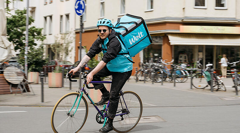 Ein Kurier auf einem Fahrrad tr&auml;gt eine gro&szlig;e, blaue Wolt-Liefertasche. Er f&auml;hrt durch eine st&auml;dtische Umgebung mit mehreren geparkten Fahrr&auml;dern im Hintergrund.