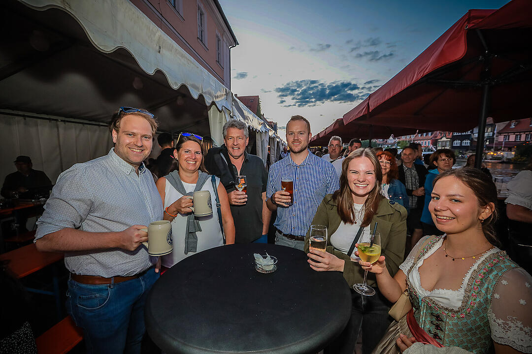 Menschen beim fröhlichen Beisammensein am Abend, trinken Bier und Wein. Hintergrund zeigt eine Festzelt-Atmosphäre, die zur Bamberger Sandkerwa passt.