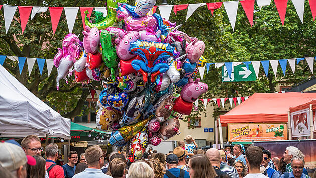Luftballons in Form von Charakteren schweben &uuml;ber einer fr&ouml;hlichen Menschenmenge bei der Bamberger Sandkerwa. Bunte Wimpel dekorieren den Platz, umgeben von St&auml;nden.