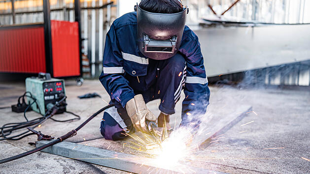Schwei&szlig;en, m&auml;nnlicher Arbeiter schwei&szlig;t Metallwerkst&uuml;cke au&szlig;erhalb der Fabrik, Schutzkleidung, Schutzhandschuhe und Schutzhelm, Schwei&szlig;funken und Licht