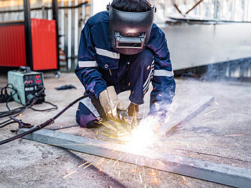 Schweißen, männlicher Arbeiter schweißt Metallwerkstücke außerhalb der Fabrik, Schutzkleidung, Schutzhandschuhe und Schutzhelm, Schweißfunken und Licht