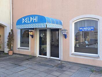 Das Bild zeigt den Eingang des griechischen Restaurants "Delphi" mit blauem Vordach. Ein Schild kündigt die Neueröffnung im Mai 2025 an.