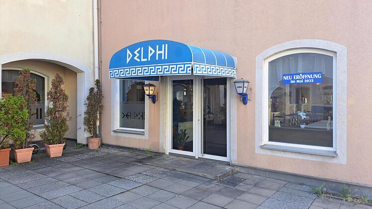Das Bild zeigt den Eingang des griechischen Restaurants "Delphi" mit blauem Vordach. Ein Schild kündigt die Neueröffnung im Mai 2025 an.