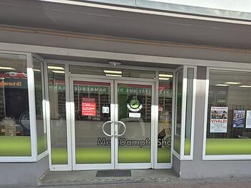 Die Fassade des geschlossenen "MeinDampferShop" in Bamberg, mit auff&auml;lligen gr&uuml;nen Akzenten auf der Glasfront, wirkt verlassen und zeigt Schilder und Poster in den Fenstern.