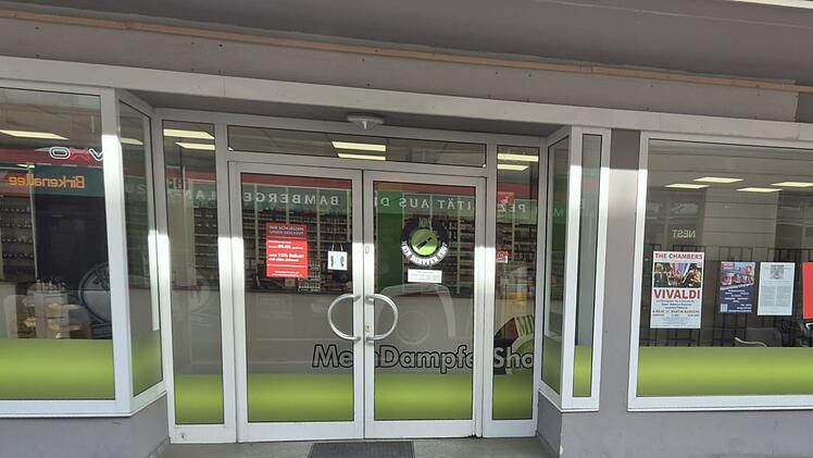 Die Fassade des geschlossenen "MeinDampferShop" in Bamberg, mit auff&auml;lligen gr&uuml;nen Akzenten auf der Glasfront, wirkt verlassen und zeigt Schilder und Poster in den Fenstern.