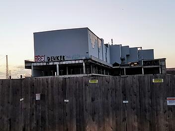 Ein teilweise erbautes Geb&auml;ude hinter einem Holzzaun. Graffiti auf der Fassade. Baustelle am fr&uuml;hen Abend.