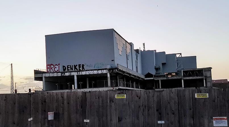 Ein teilweise erbautes Geb&auml;ude hinter einem Holzzaun. Graffiti auf der Fassade. Baustelle am fr&uuml;hen Abend.