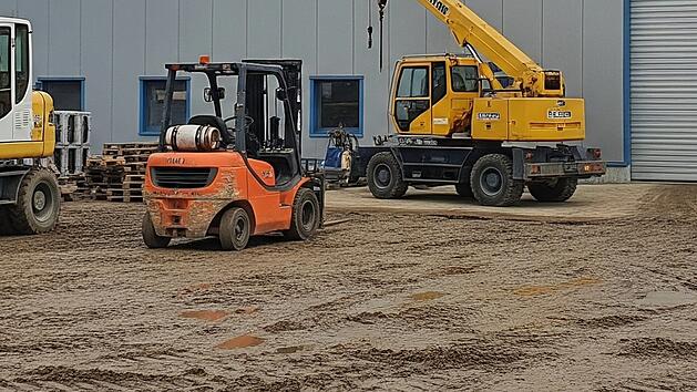 Firmensitz einer mittelständischen Baufirma. Vor einer Halle sind ein Bagger, ein Gabelstapler und ein kleiner Kran zu sehen.