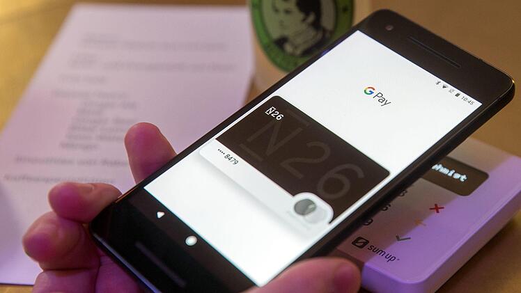 Ein Gast demonstriert mit einem Smartphone und der App des neuen Bezahldienstes Google Pay in einem Cafe das Bezahlen mit dem Handy. Bei dem Dienst ersetzt ein Telefon mit dem Google-Betriebssystem Android die Kreditkarte, zum Zahlen hält man das Gerät im Laden an das Terminal. Die Kassentechnik muss dafür kontaktloses Bezahlen unterstützen - ein großer Teil der Terminals in Deutschland wurde bereits entsprechend umgerüstet. Foto: Jens Büttner/dpa