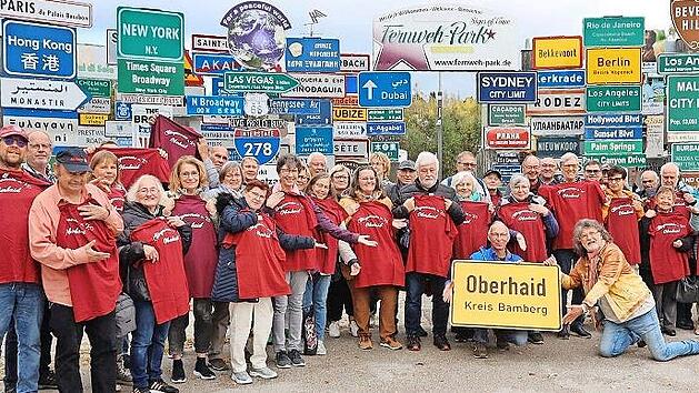 Die Ausflugsgruppe des B&uuml;rgervereins Oberhaid mit Werner Hoffmann (hinter dem Ortsschild) mit Fernweh-Park Initiator Klaus Beer (knieend) bei der Pr&auml;sentation des Ortsschildes vor der Panorama-Schilder-Weltkulisse des Fernweh-Parks.