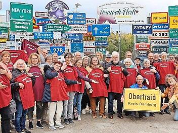 Die Ausflugsgruppe des Bürgervereins Oberhaid mit Werner Hoffmann (hinter dem Ortsschild) mit Fernweh-Park Initiator Klaus Beer (knieend) bei der Präsentation des Ortsschildes vor der Panorama-Schilder-Weltkulisse des Fernweh-Parks.