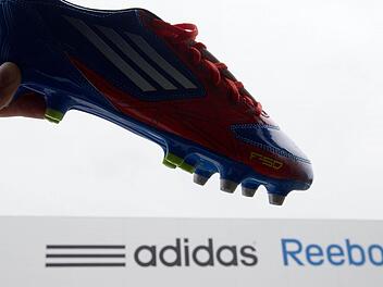 Ein Adidas-Schuh wird am 03.07.2013 in Rieste (Niedersachsen) vor das Logo von Adidas und Reebok gehalten. Foto: Friso Gentsch/dpa