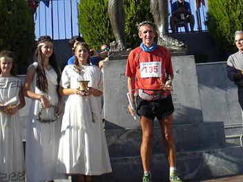 Ziel des Spartathlons ist keine Linie. Für die Teilnehmer ist der Lauf dann abgeschlossen, wenn sie den Fuß der Statue des legendären Königs der Spartaner, Leonidas, berühren. Davor steht Hubert Karl mit Lorbeerkranz und Urkunde, daneben die Ehren-Delegation der Stadt Sparta im historischen Gewand.