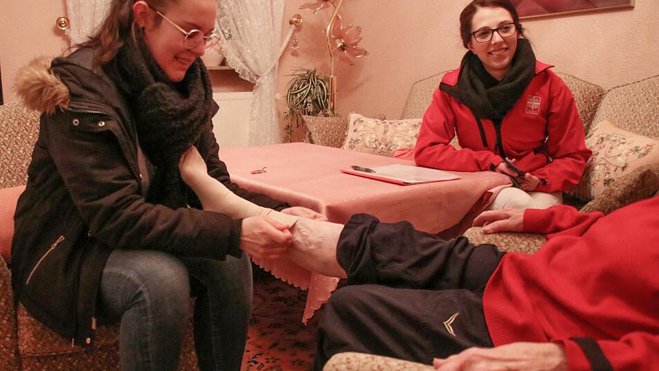 "Einen fröhlichen Menschen hat Gott lieb", erklärt die Seniorin ihren Pflegerinnen, als Michelle Köhne ihr Thrombosestrümpfe anzieht. Katharina Merkel kümmert sich um die Dokumentation.  Foto: Sebastian Schanz
