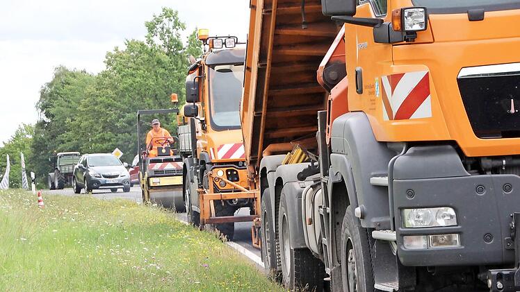 Eine kleine Karawane orangefarbener Fahrzeuge vom Straßenbau erledigt die Arbeiten. Die Walze am Ende verdichtet den frischen Asphaltauftrag, so dass die Straße sogleich wieder befahren werden kann.
