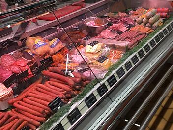 Eine Metzgereitheke mit verschiedenen Fleisch- und Wurstsorten, beschriftet mit Preisschildern. Die Auslage wirkt gut gefüllt und vielfarbig arrangiert.